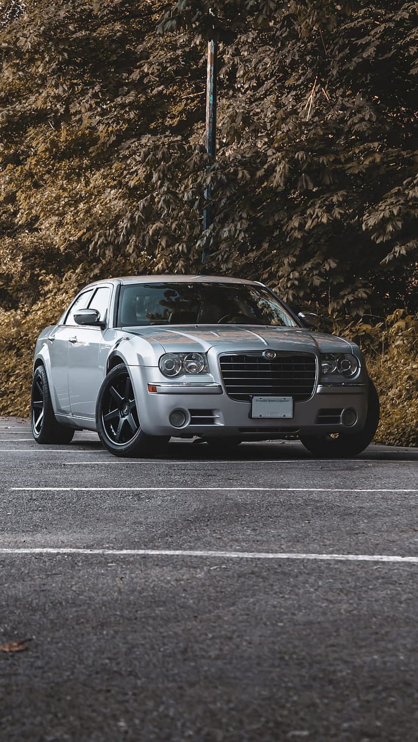 Silver Chrysler 300 sedan parked on an asphalt lot with custom black rims and a backdrop of dense forest trees.