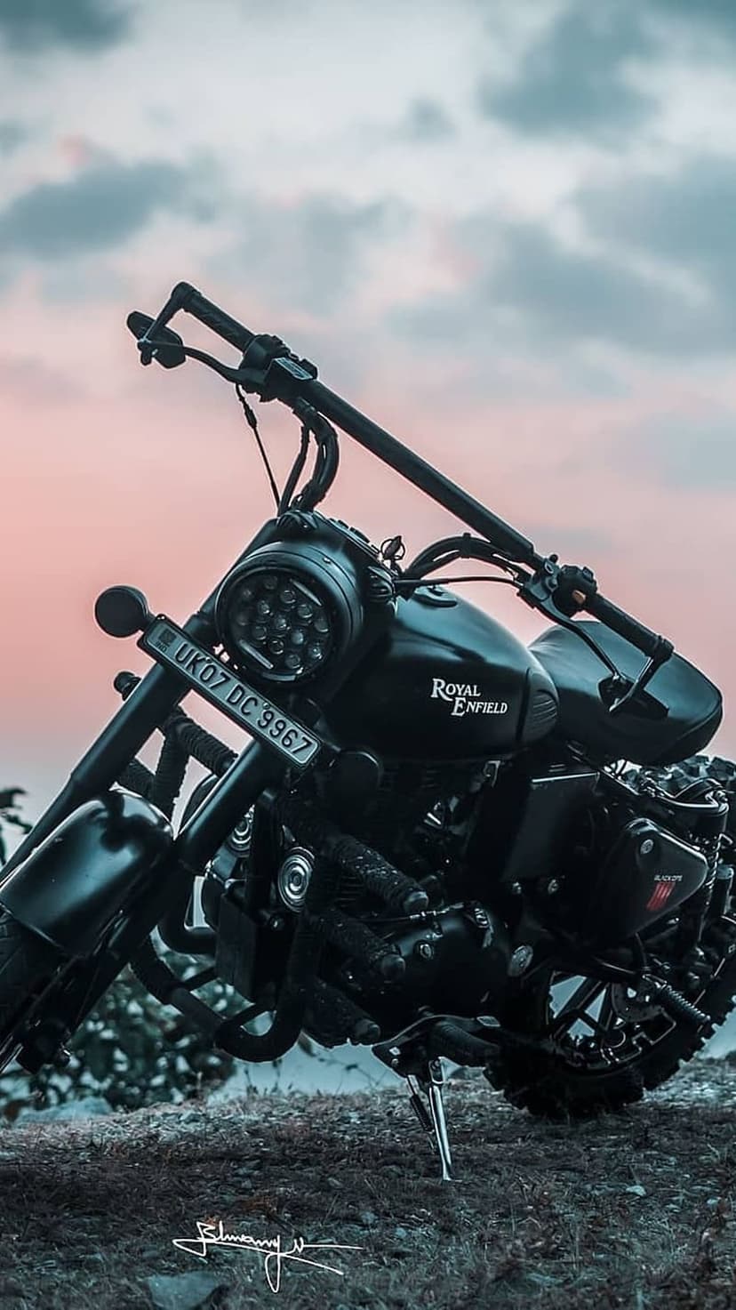 Matte black Royal Enfield motorcycle parked on a rocky slope against a vibrant orange and purple sunset sky.