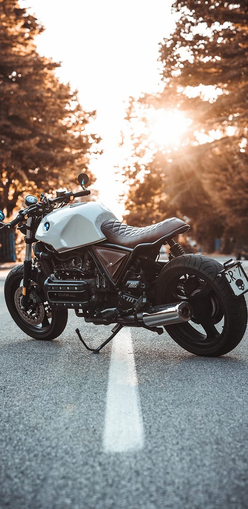 Custom BMW motorcycle with a brown seat and chrome exhaust sits on an asphalt road during a golden hour sunset.
