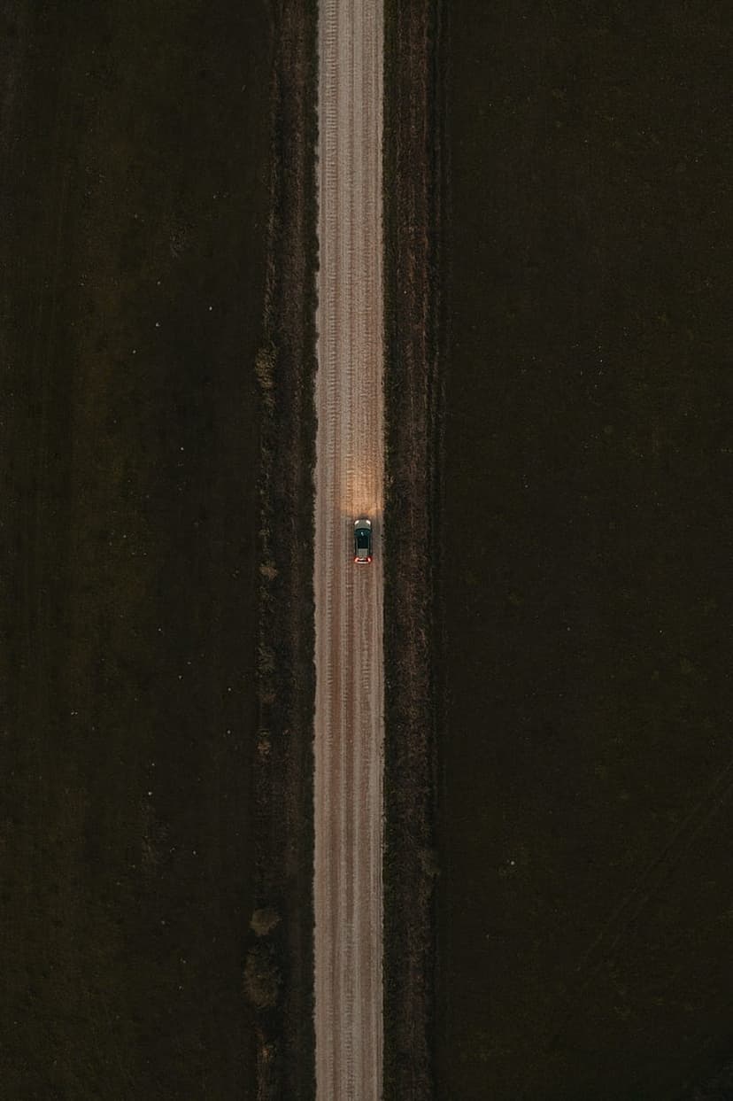 White headlights of a car illuminate a dusty brown road from an overhead drone perspective in a dark landscape.