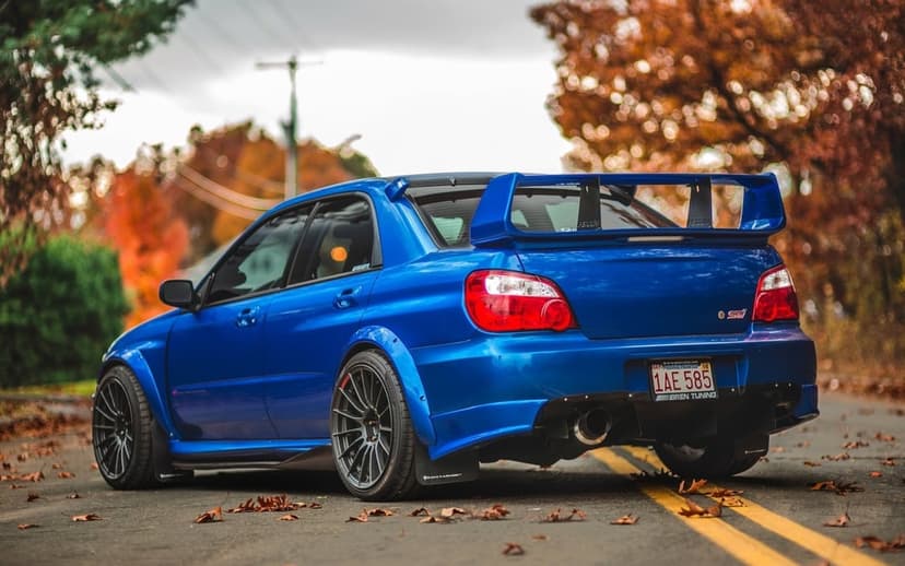Blue Subaru WRX STI widebody with a large rear wing parked on a road covered in vibrant orange autumn leaves.