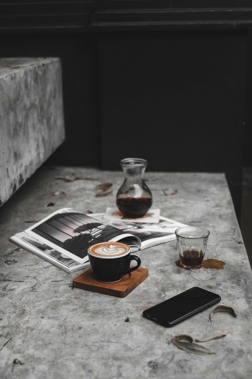 Ceramic mug with latte art on a wooden coaster beside an open magazine and smartphone on a concrete table.