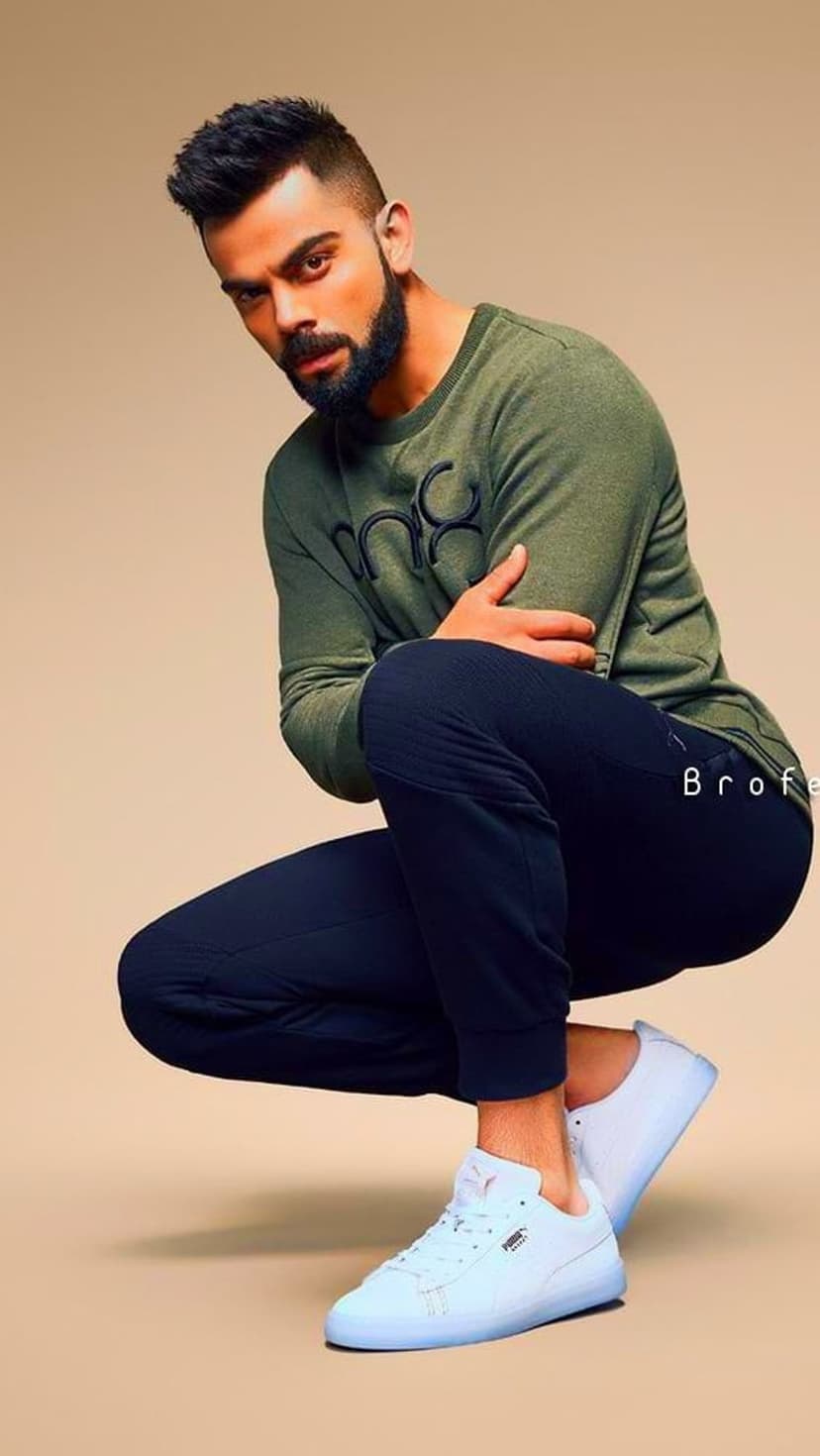 Virat Kohli wearing a green sweatshirt and navy pants while crouching in white sneakers against a grey backdrop.