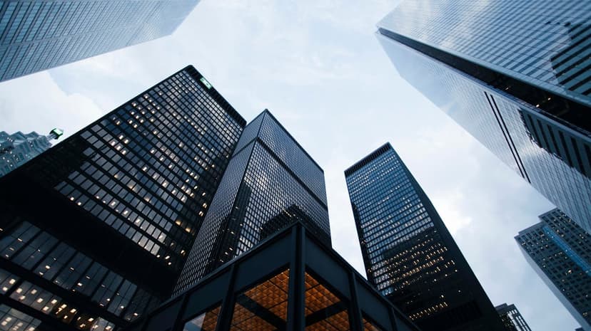Glass skyscrapers rise into a gray sky from a low angle, reflecting the surrounding city and clouds.