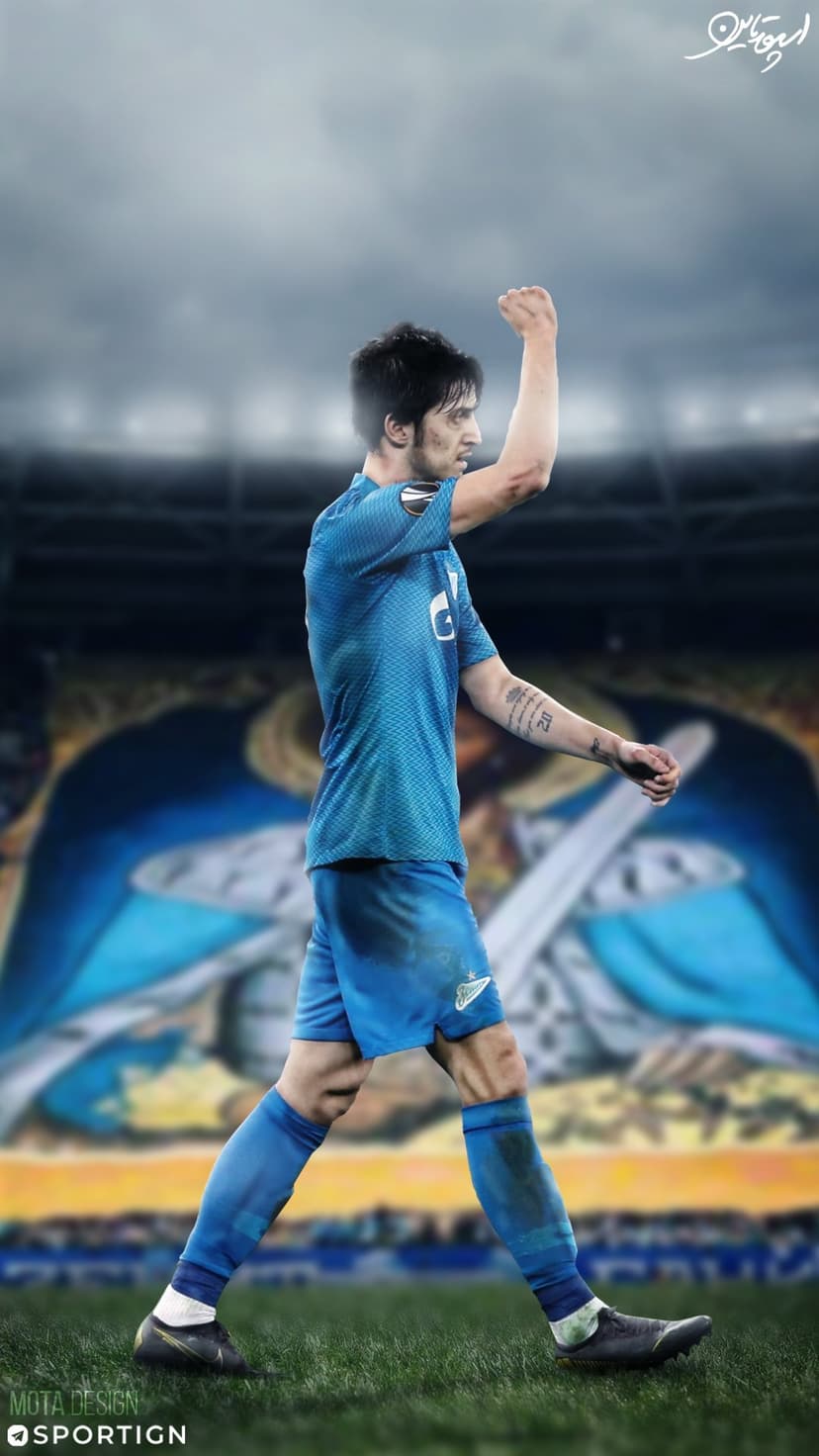 Football player in a blue kit raises a fist in triumph on a green field before a blurred stadium crowd.