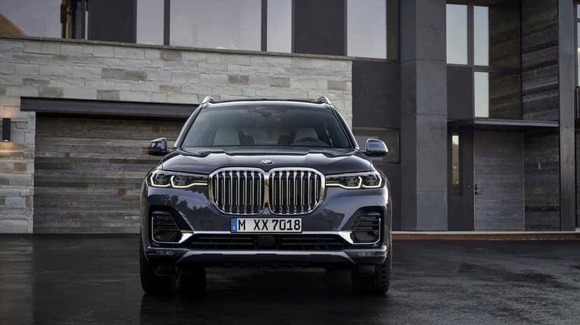 Front view of a dark grey BMW X7 SUV parked on a wet driveway in front of a modern stone house with large windows.
