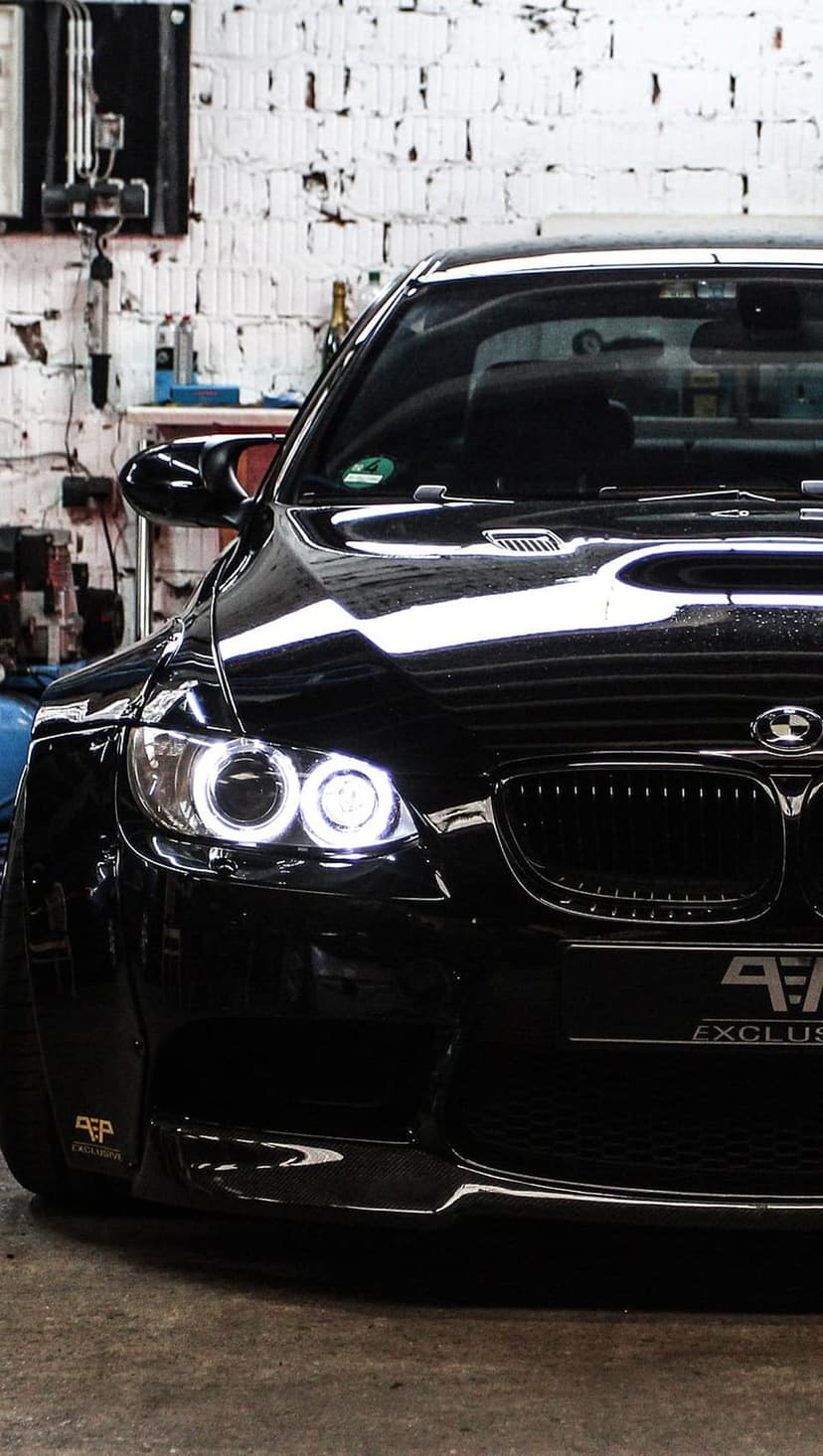 Black BMW M3 with glowing halo headlights parked in a clean workshop against a white brick and tool wall.