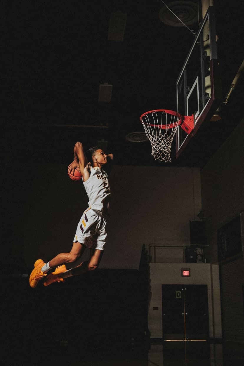 Basketball player in a white and yellow jersey mid-dunk against a dark gym background with dramatic lighting.