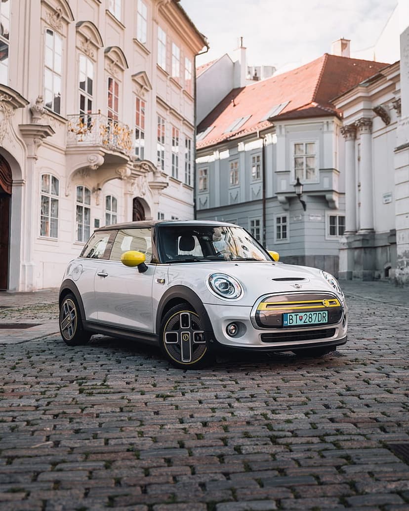 White Mini Electric car with yellow accents parked on a cobblestone street lined with historic urban buildings.