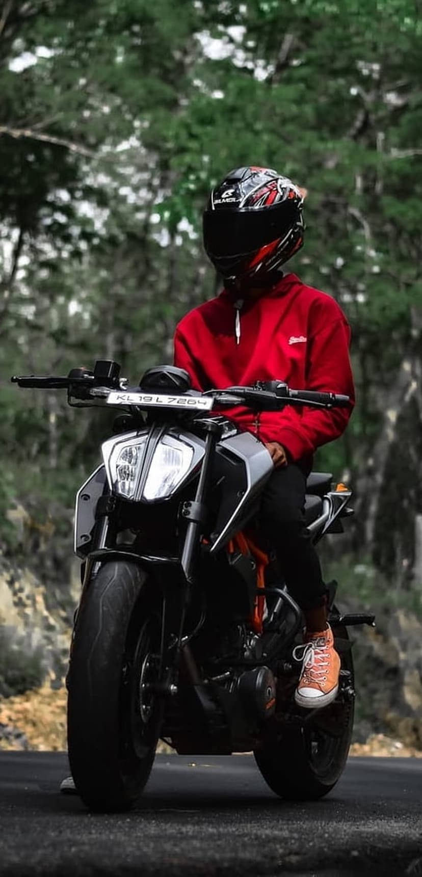 Biker in a red hoodie and helmet on a KTM motorcycle surrounded by green trees in a dense forest environment.