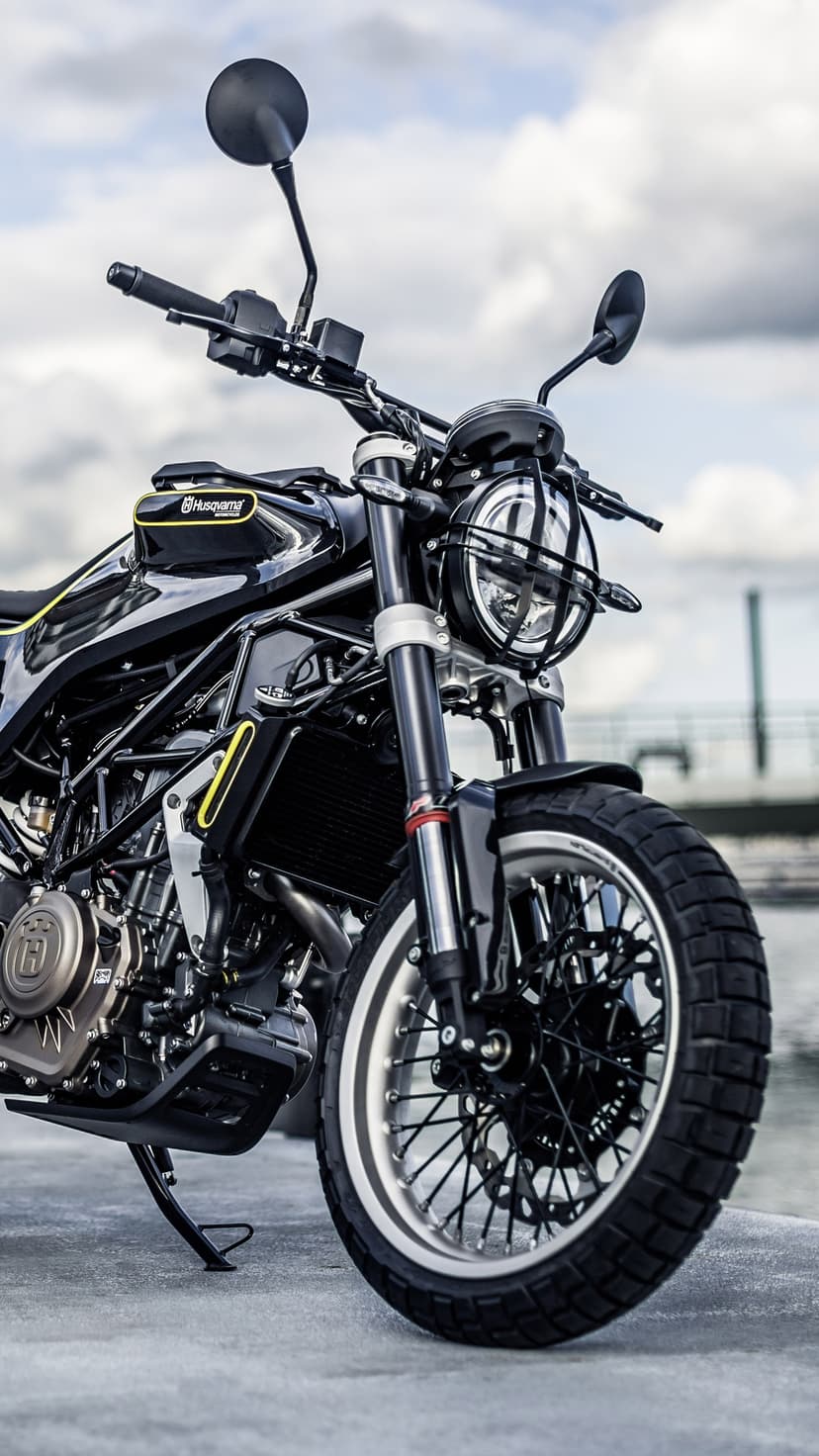 Black Husqvarna Vitpilen motorcycle with a round headlight grille parked on concrete near water and cloudy sky.