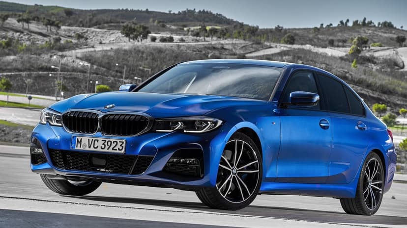 Blue BMW 3 Series sedan parked on a grey road with a soft-focus hill background under bright daylight.