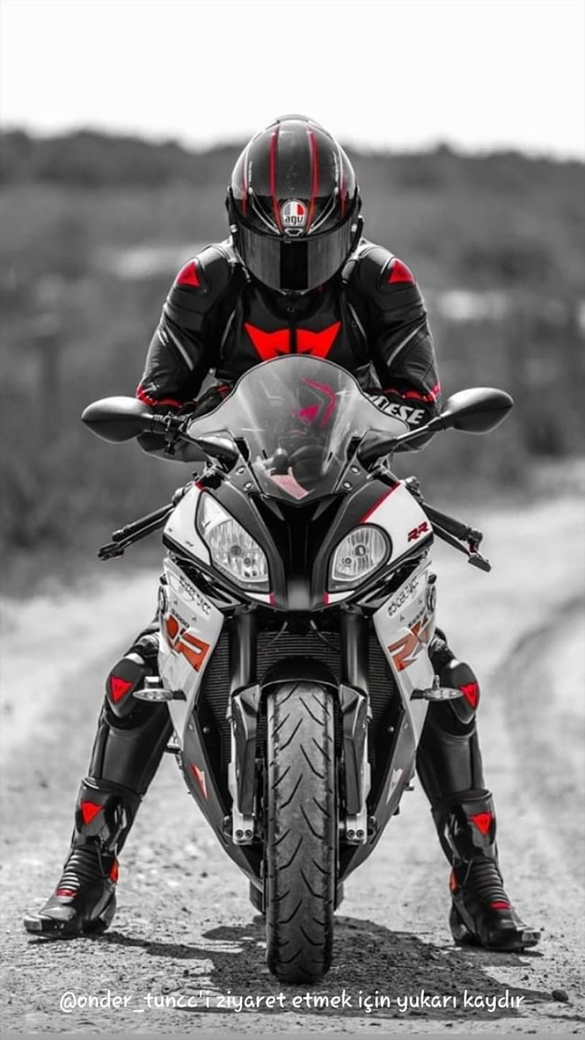 Motorcyclist in a black AGV helmet and red racing suit sits on a white sportbike over a gravel path background.