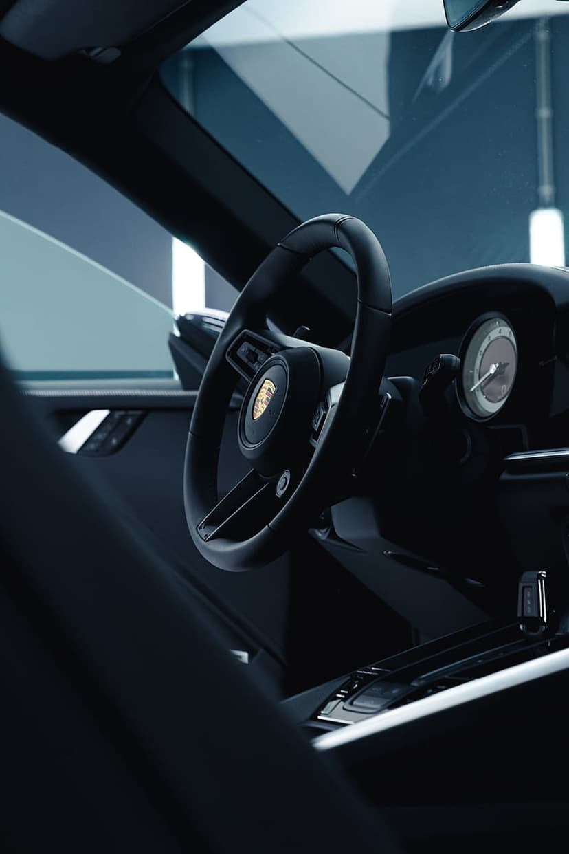 Porsche steering wheel with a gold crest and an analog dashboard dial inside a dark, leather luxury car cabin.