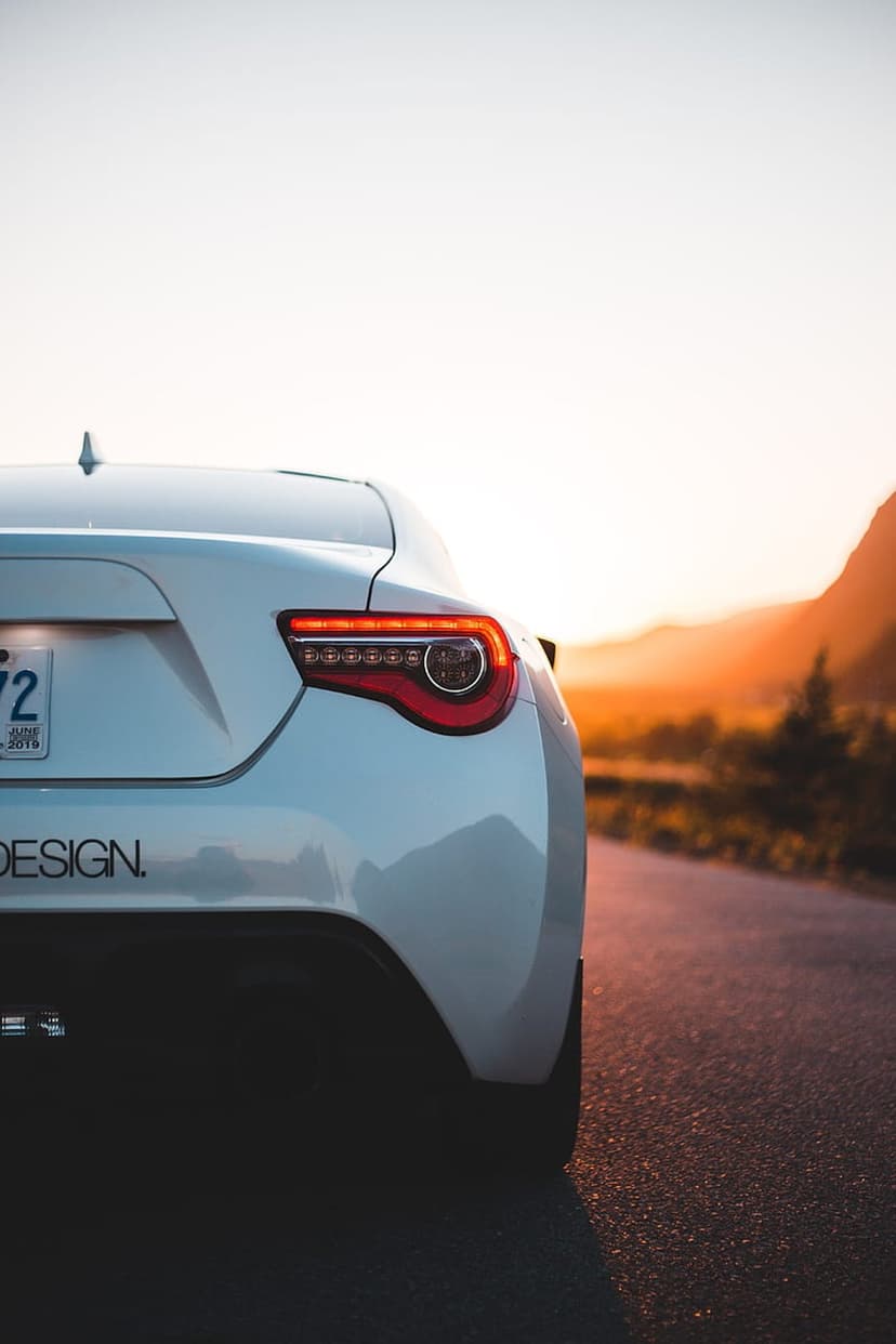 White sports car tail light glowing red on a mountain road under a vibrant orange and yellow sunset sky.