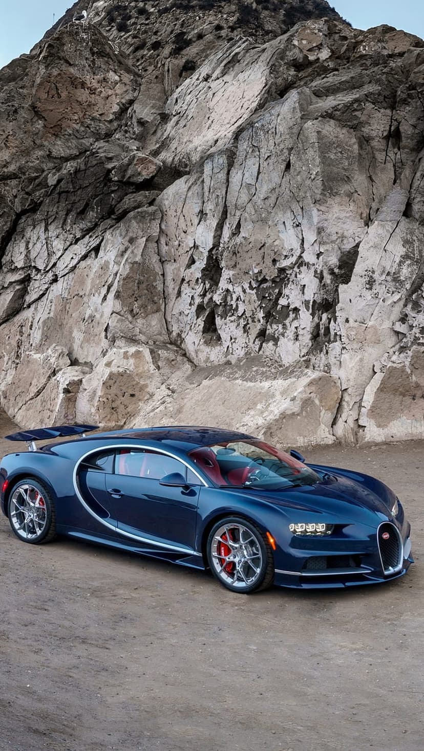 Blue Bugatti Chiron parked on a rocky coastal road with a red interior, silver wheels, and a steep cliff backdrop.