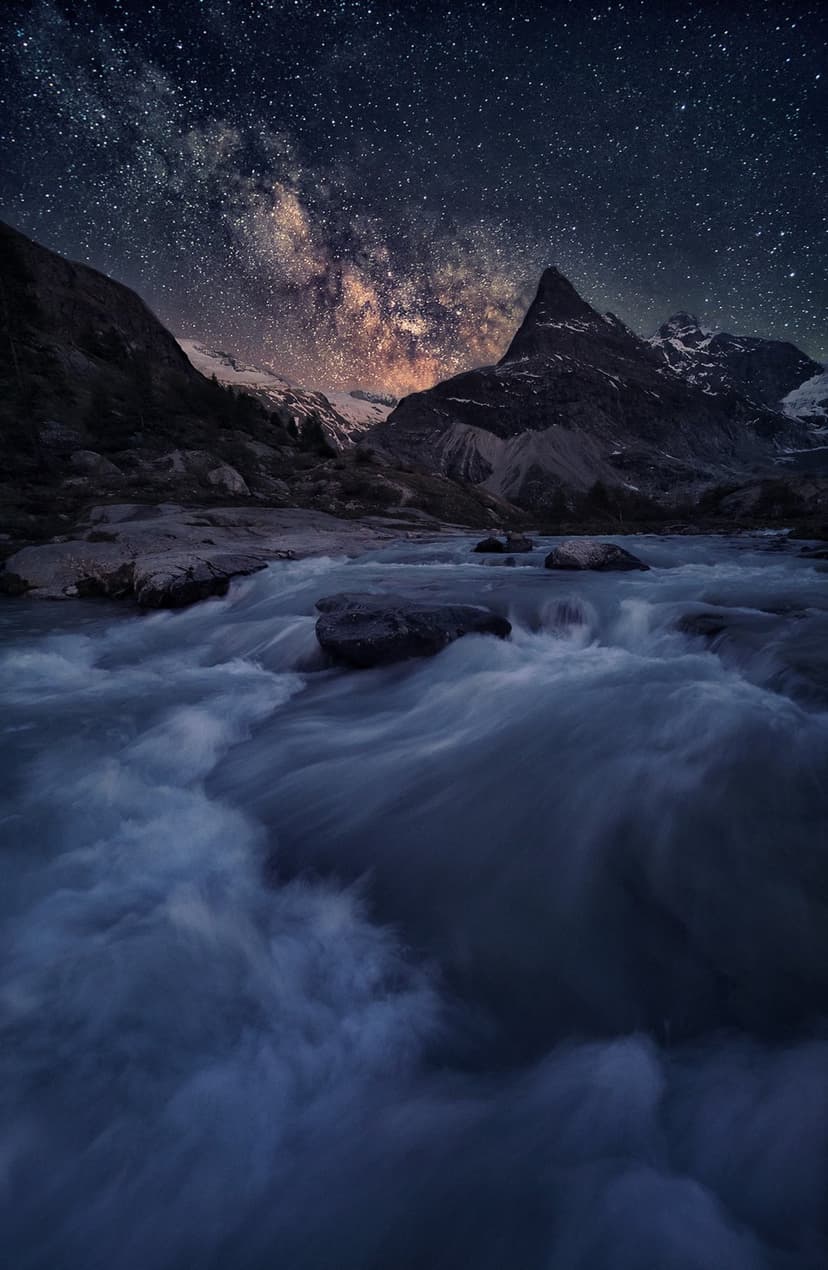 Milky Way galaxy arches over snow-capped mountains and a rushing river with motion blur in a dark night sky.