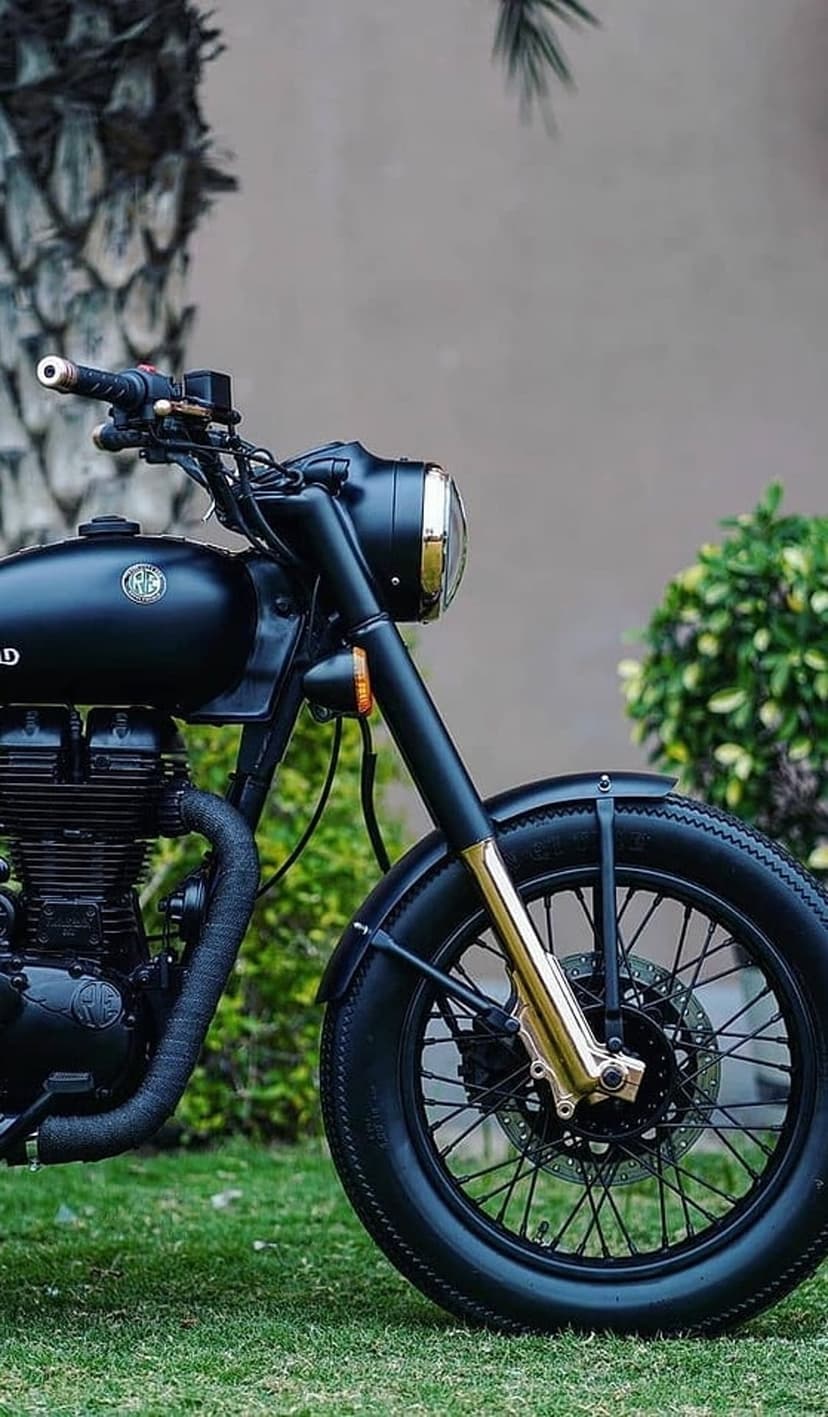 Royal Enfield motorcycle in matte black with gold forks parked on a green lawn showing engine and wheel details.
