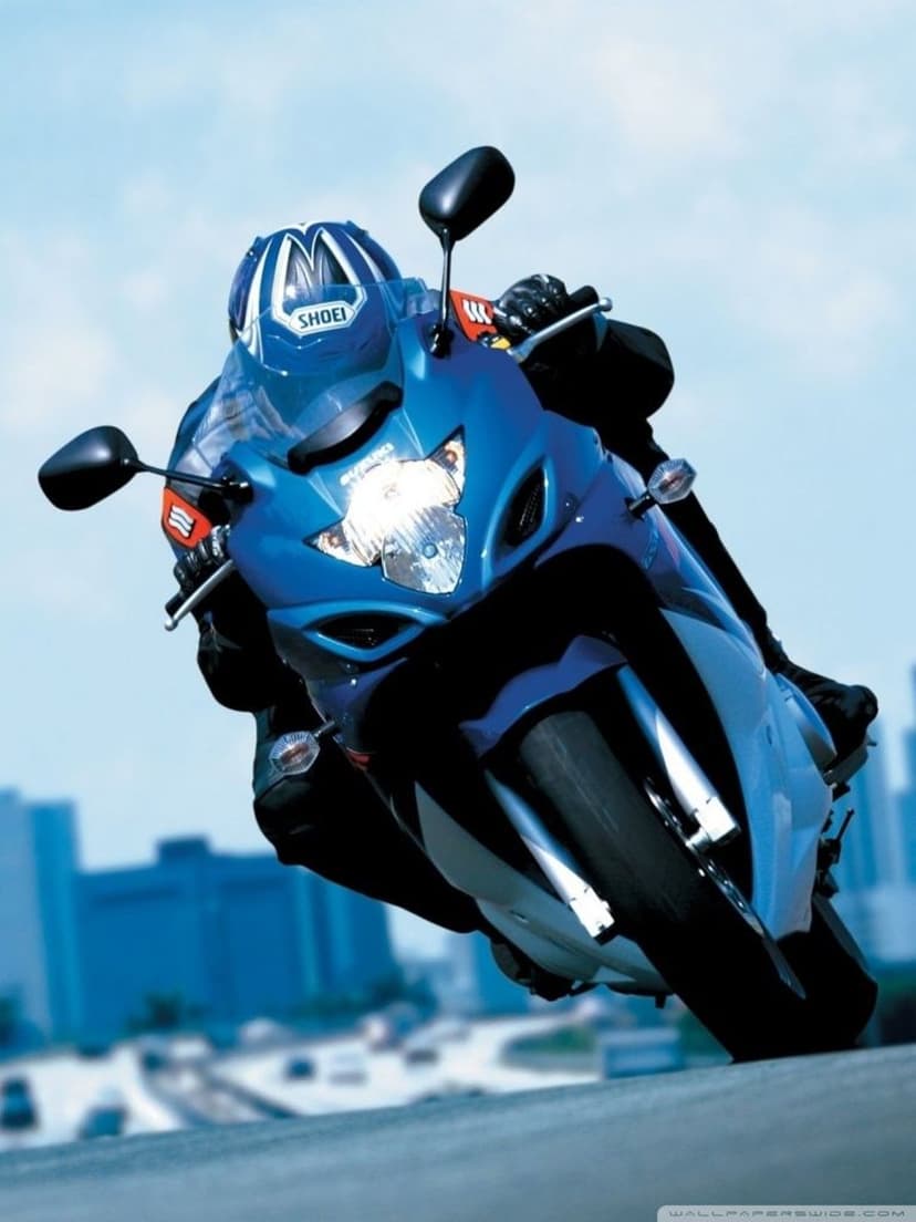 Blue sport motorcycle rider in a racing suit leans into a turn against a blurred cityscape and sky.