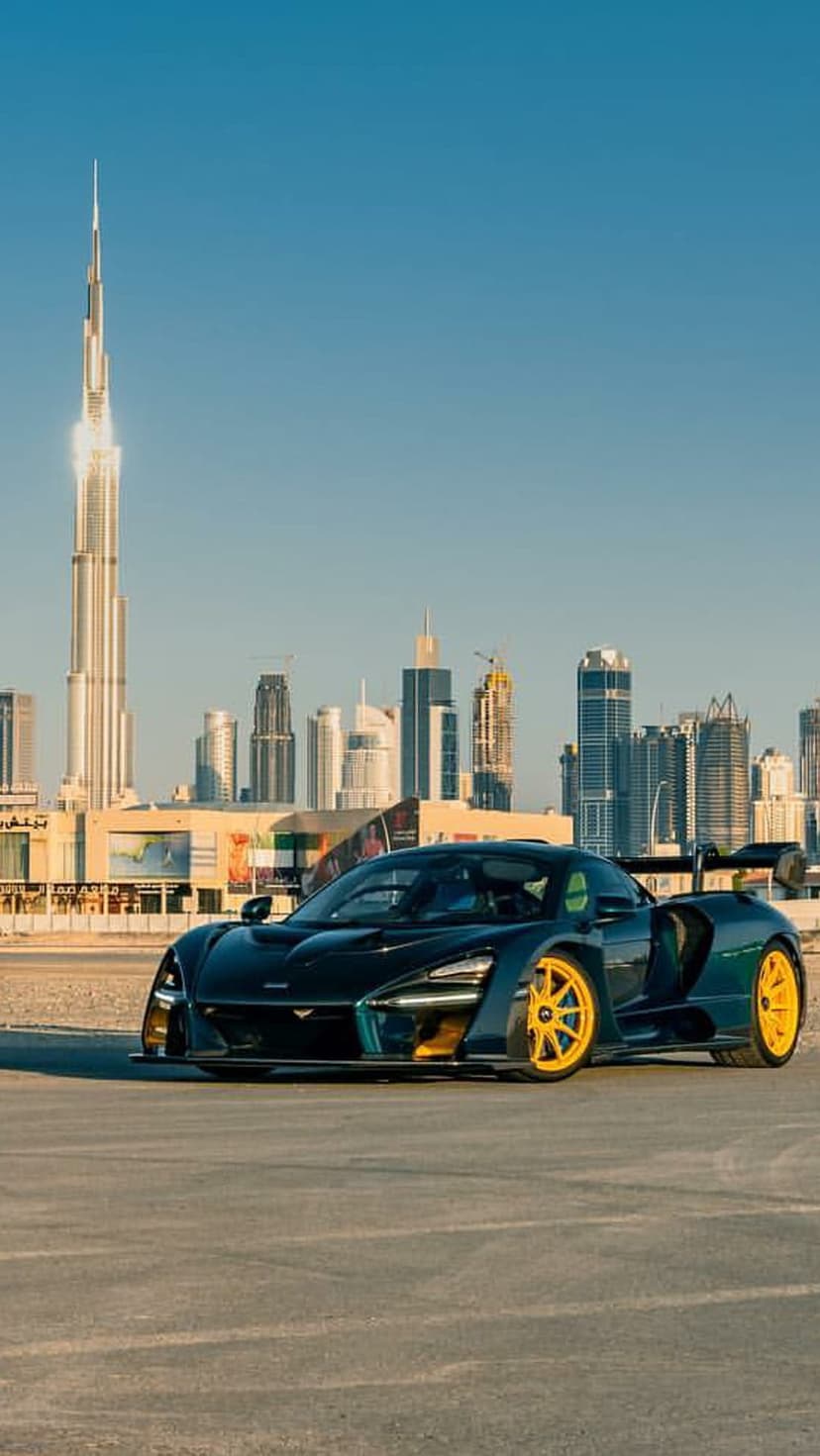 Dark blue McLaren Senna with yellow wheels parked on an urban road with the Burj Khalifa towering in the distance.