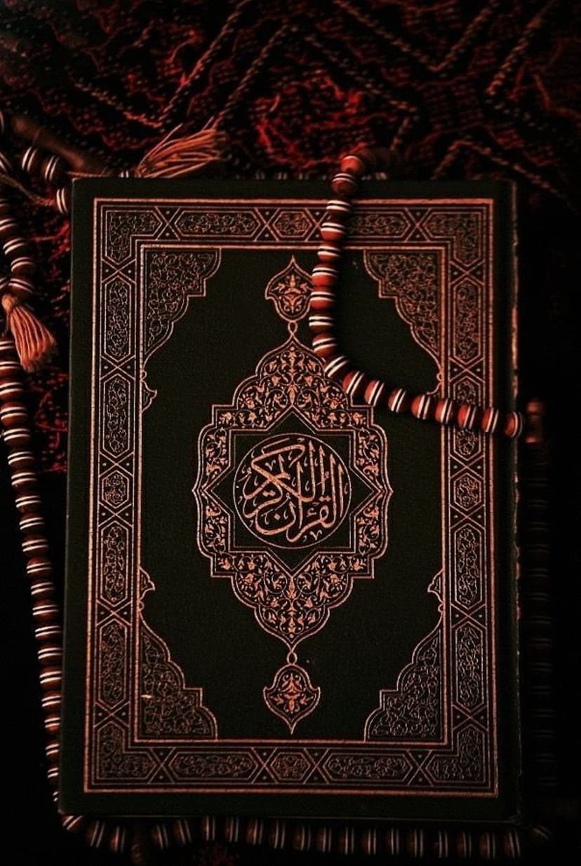 Green Quran with gold Arabic calligraphy and red prayer beads on dark textured cloth in a vertical close-up shot.