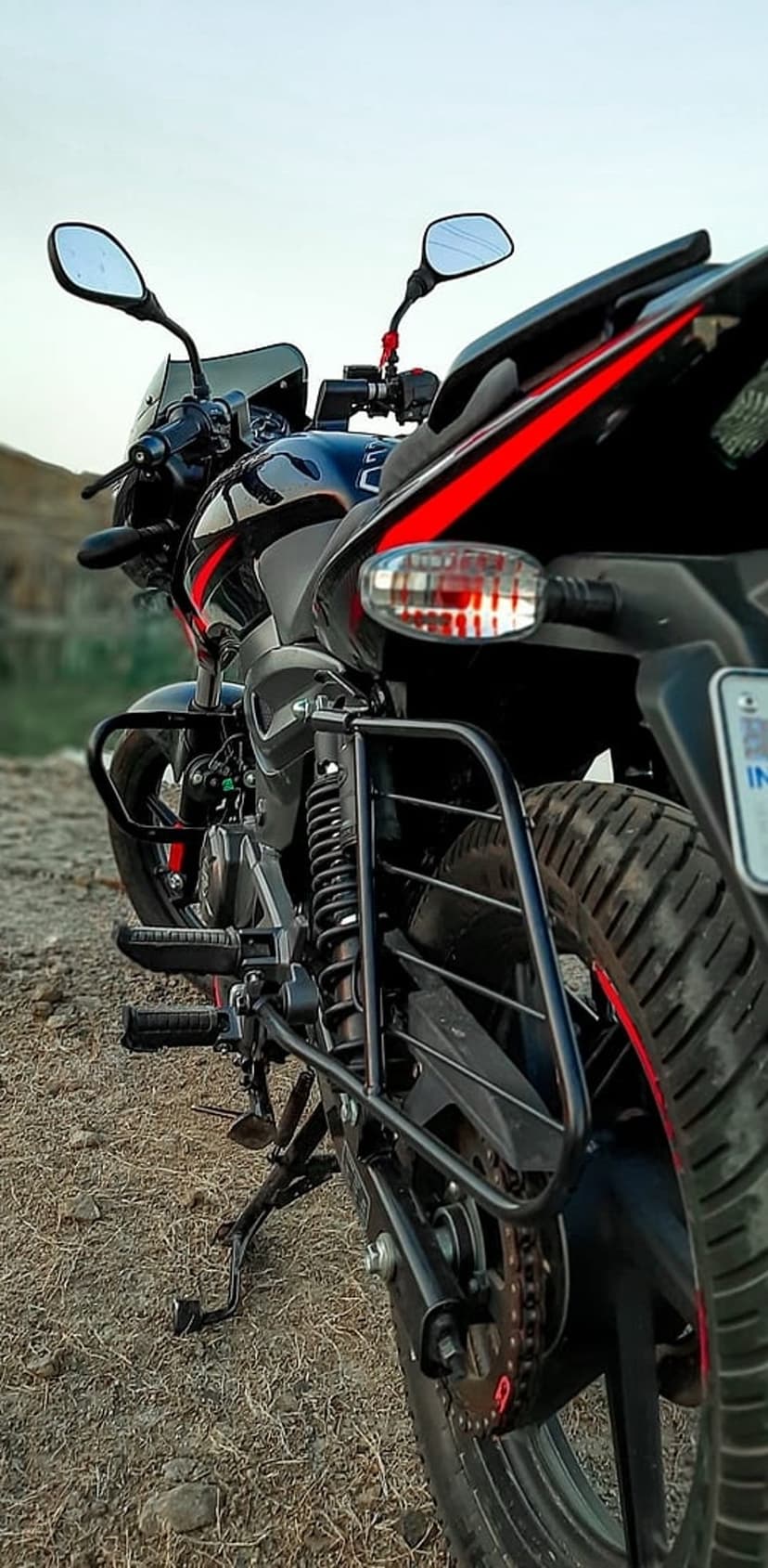 Black Bajaj Pulsar motorcycle featuring red accents parked on gravel with a blurred nature background.