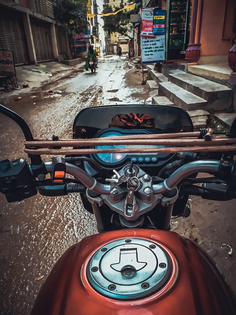 Motorcycle handlebars carrying timber navigate a wet, narrow street lined with buildings during a rainy day.