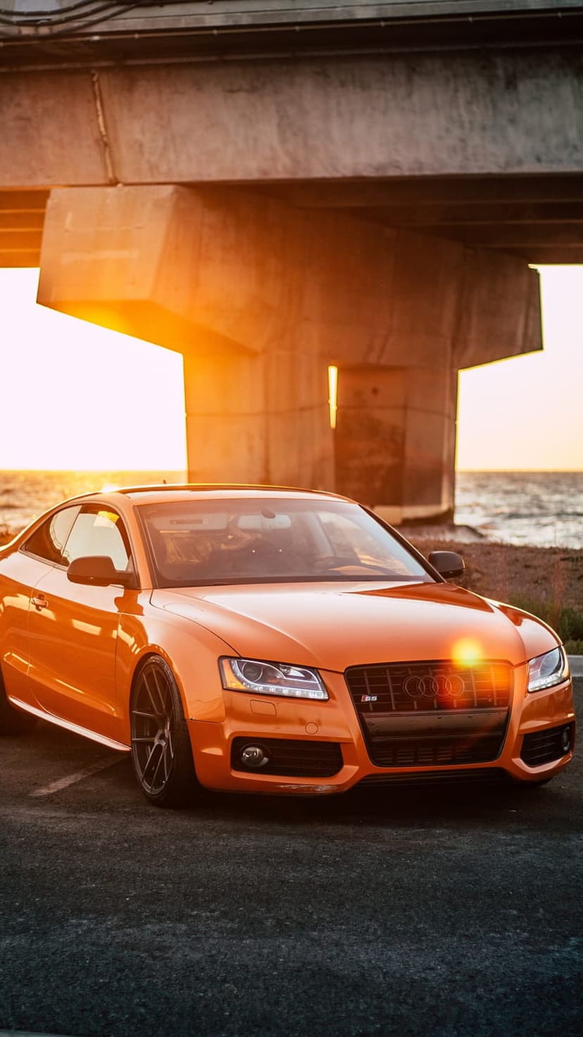 Orange Audi S5 Coupe parked under a concrete bridge with a golden sunset over the ocean in the background.