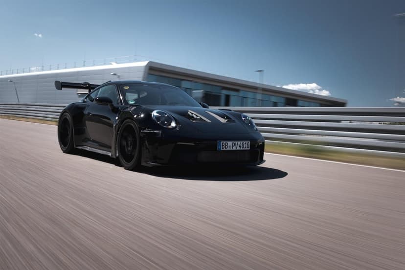 Black Porsche 911 GT3 RS prototype speeding on a track past a silver building under a clear blue sky.