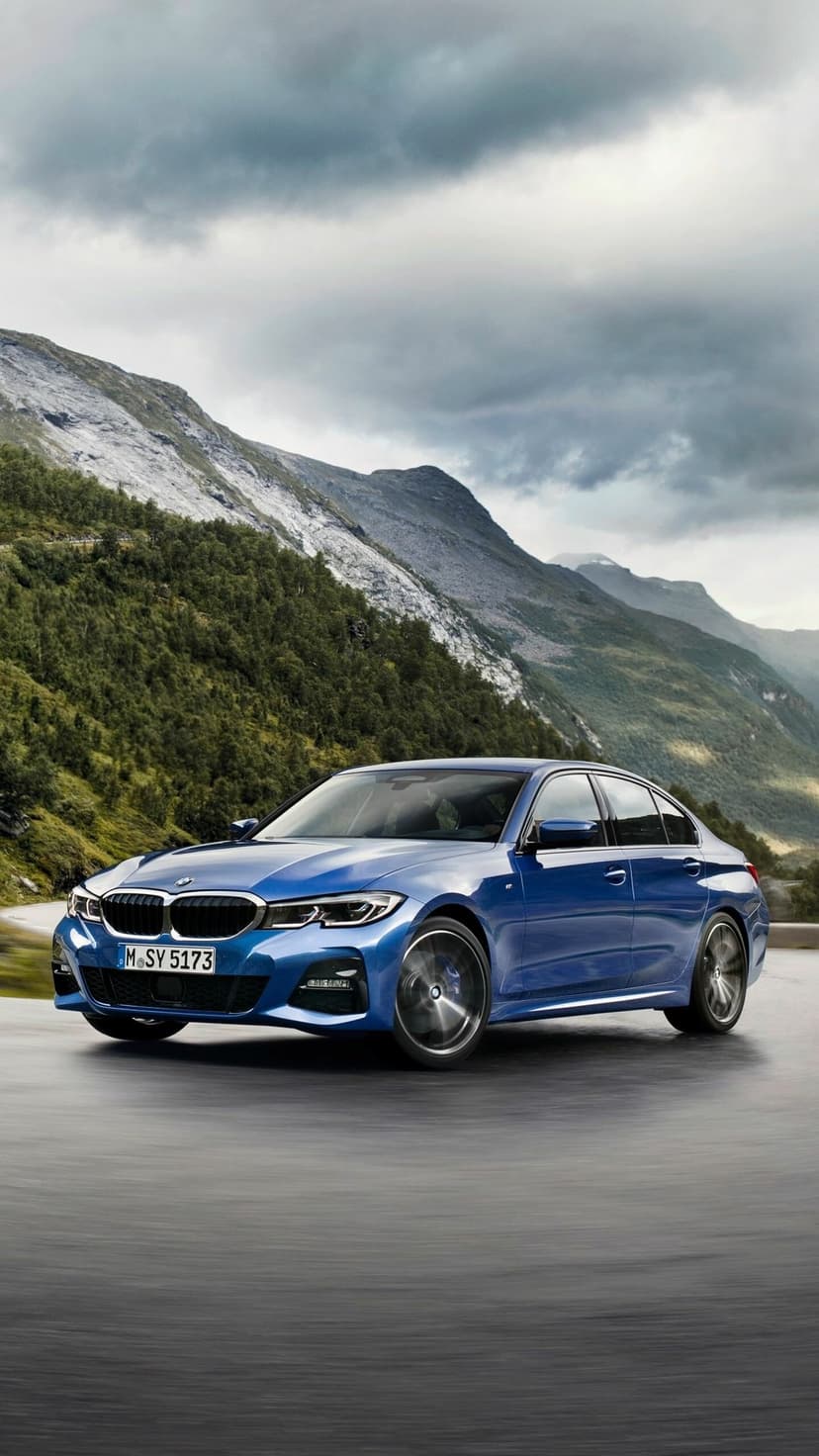 Blue luxury sedan driving on a winding mountain road under a cloudy sky with green slopes and rocky peaks.