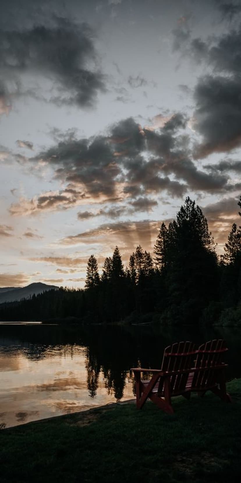 Red Adirondack chairs face a calm lake reflecting a golden sunset with distant mountains and evergreen trees.