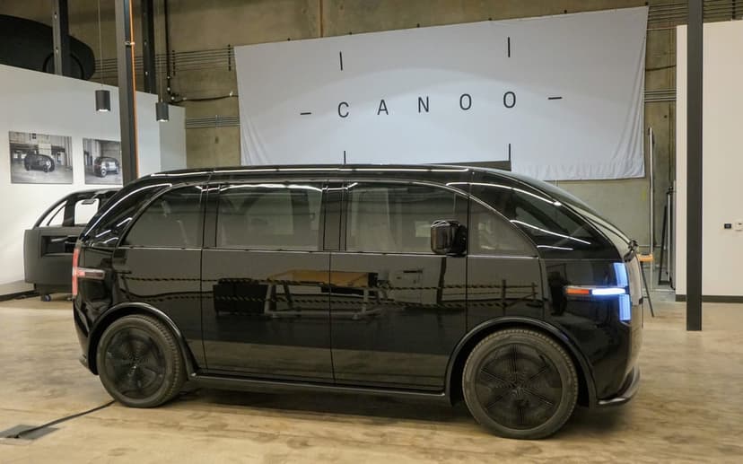 Side profile of a glossy black Canoo electric vehicle in a studio with a white logo banner and concrete floor.