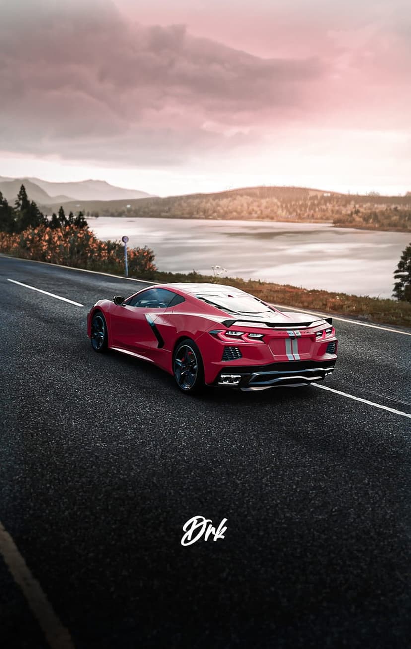 Red Corvette C8 driving on a winding asphalt road by a calm lake under a pink and purple sunset sky.