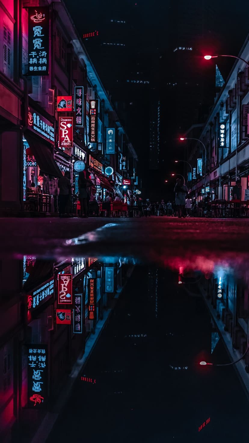 Wet city street at night reflecting red and blue neon signs with light trails on damp asphalt pavement.