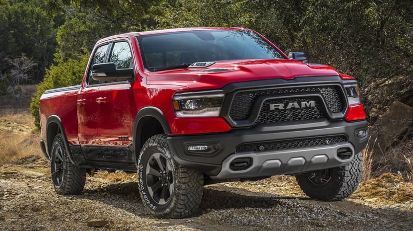 Red RAM 1500 Rebel pickup truck parked on a rocky gravel trail with lush green trees and bright daylight.