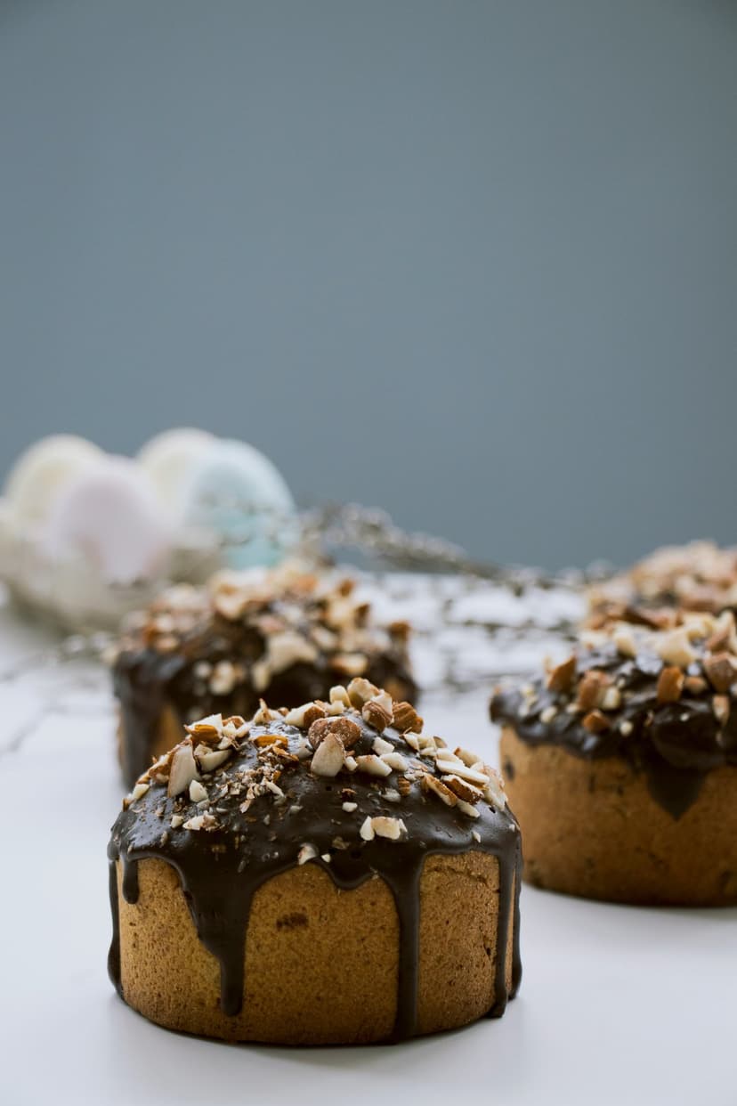 Four small chocolate-drizzled cakes with chopped almonds on a wire rack against a soft blurred background.