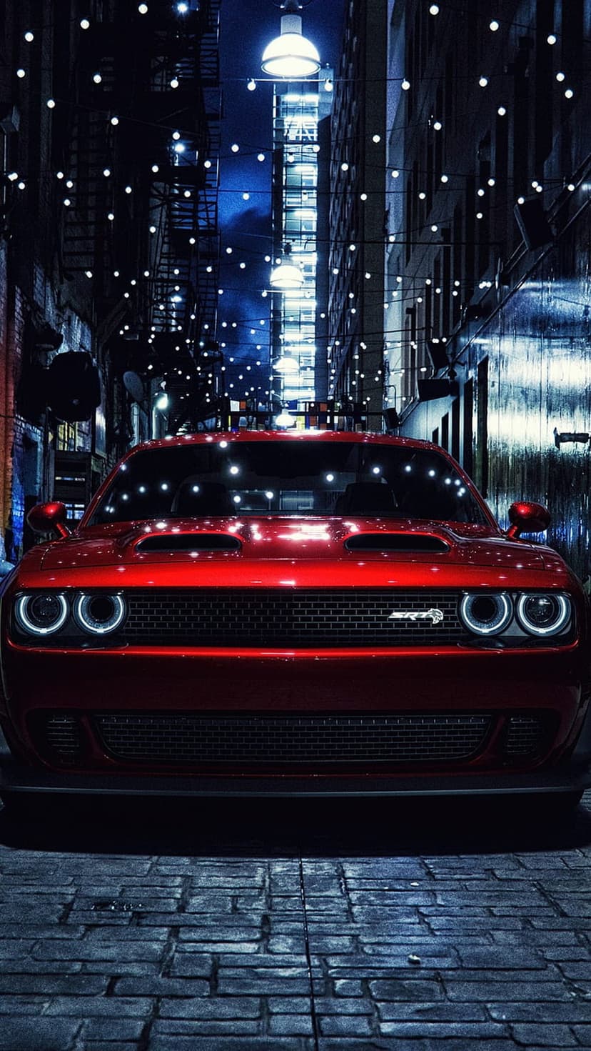 Red Dodge Challenger sits on cobblestones in a dark alley illuminated by overhead string lights and neon.