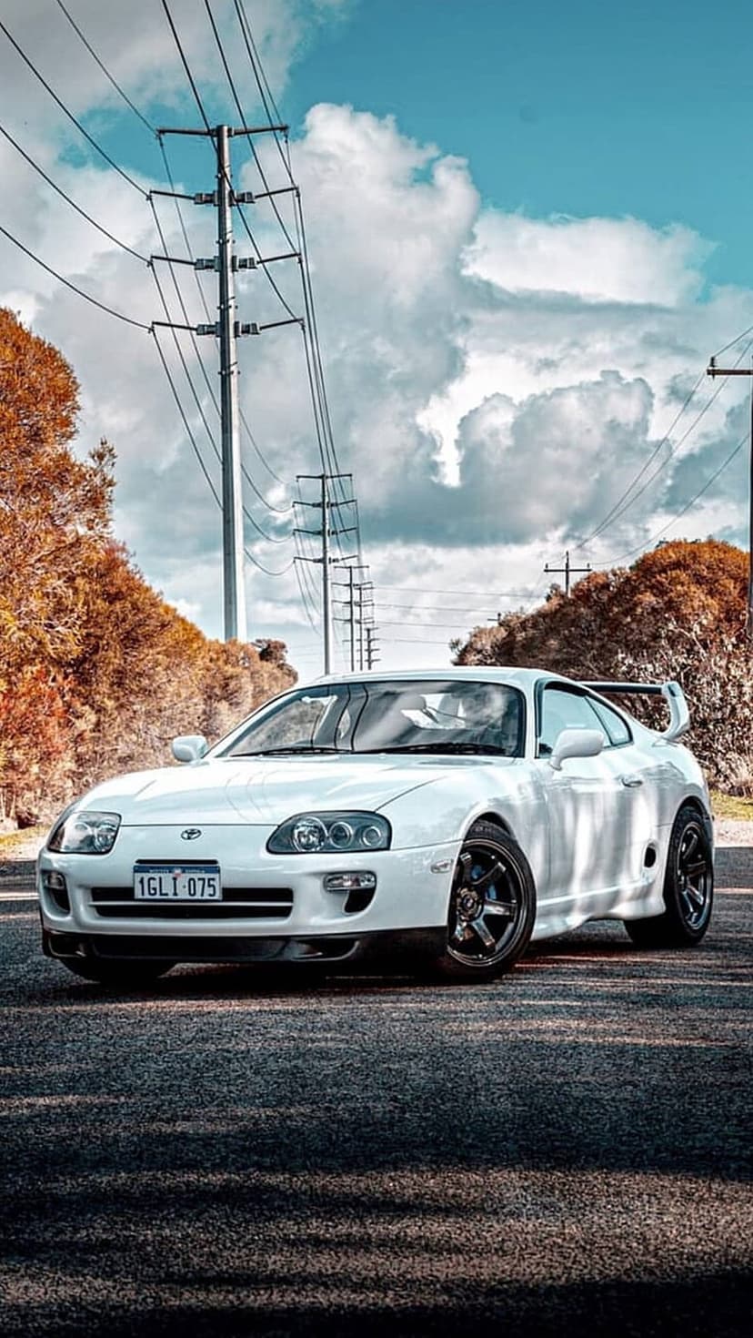 White Toyota Supra sports car parked on an asphalt street beneath industrial power lines and a cloudy blue sky.