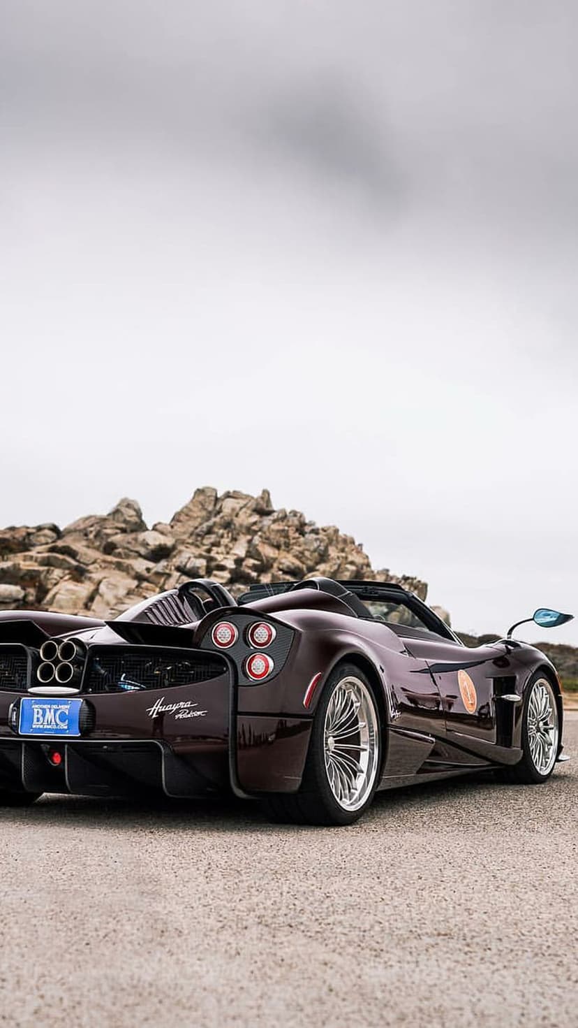 Maroon Pagani Huayra Roadster rear view with quad exhausts parked on asphalt beside a rugged rocky mountain.