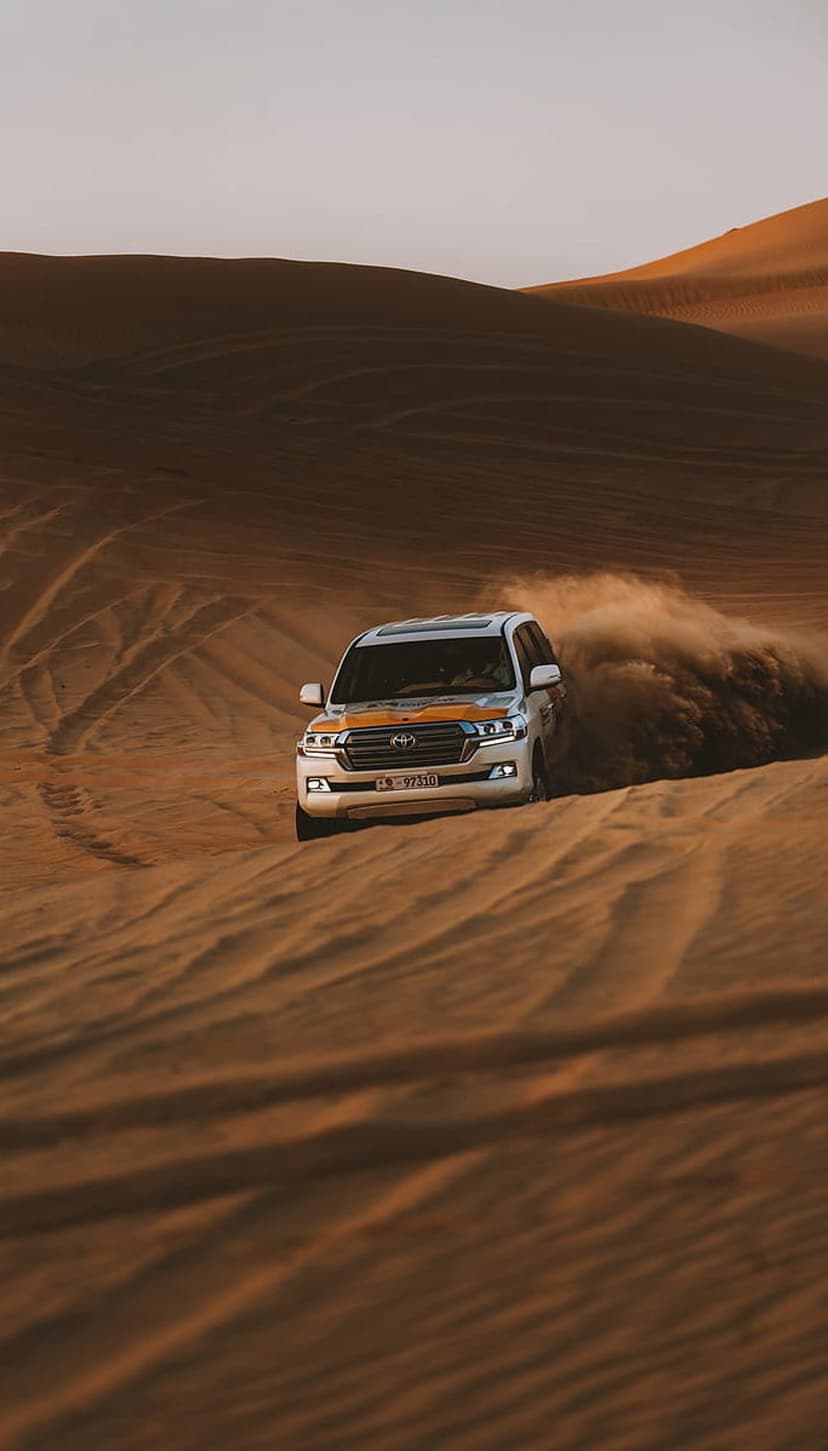 White Toyota Land Cruiser SUV kicking up a large sand cloud while driving across rolling desert dunes.