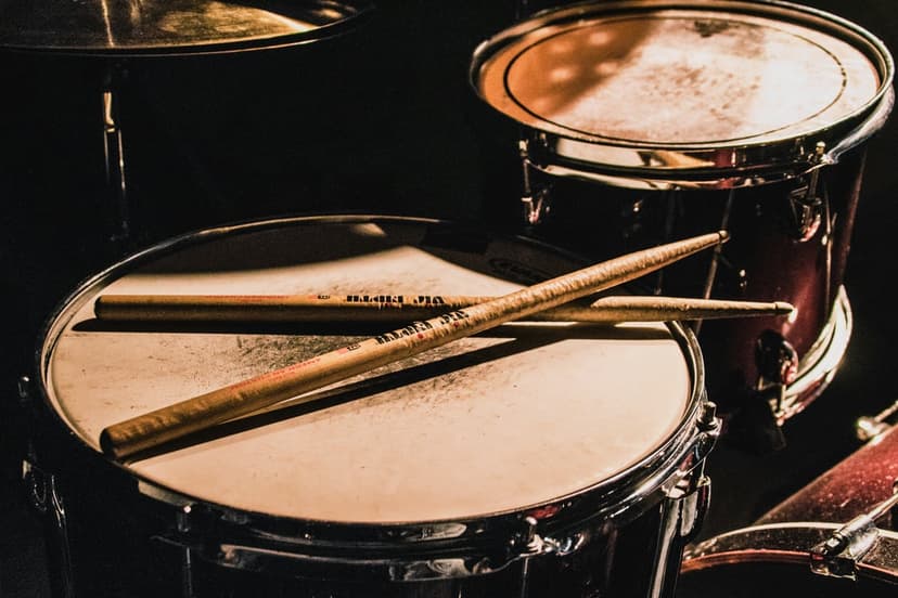 Drumsticks resting on a white snare drum head with warm golden lighting and blurred cymbals in the background.