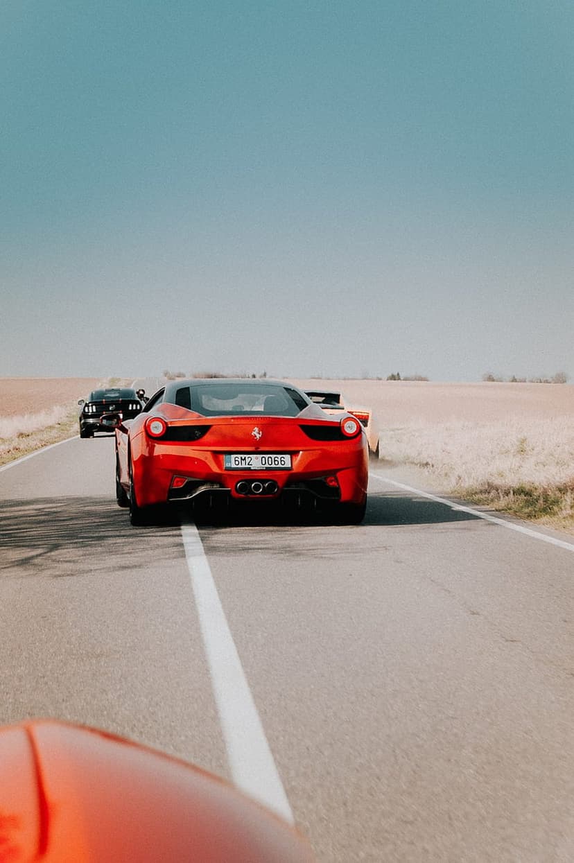 Red Ferrari leads a black Mustang on a sunlit asphalt road under a clear blue sky during a high-speed race.