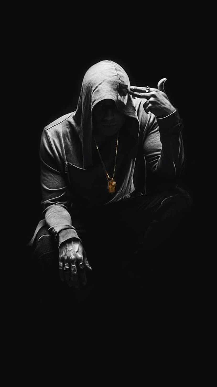Hooded figure in shadows holds a hand to their temple wearing a gold chain in a monochrome vertical portrait.