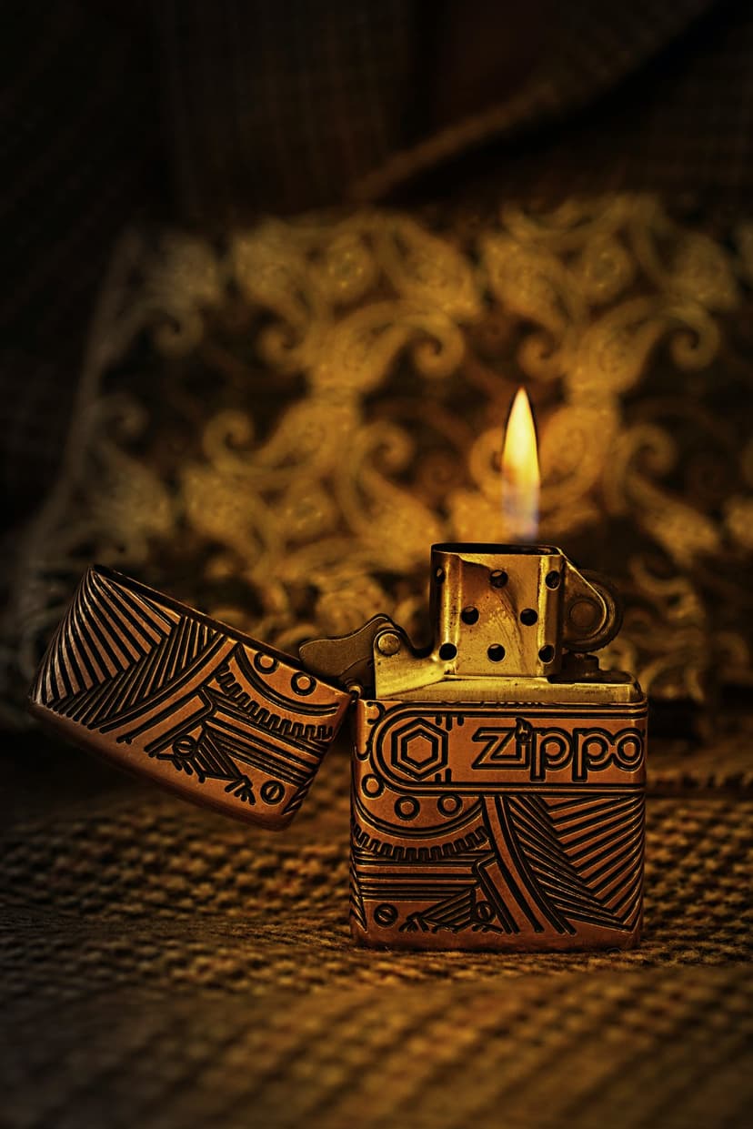 Vintage brass lighter with intricate carvings emits a bright orange flame against a dark blurred background.