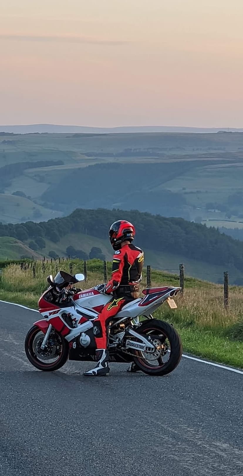 Motorcyclist in red racing gear on a Yamaha bike overlooking rolling green hills under a pink twilight sky.