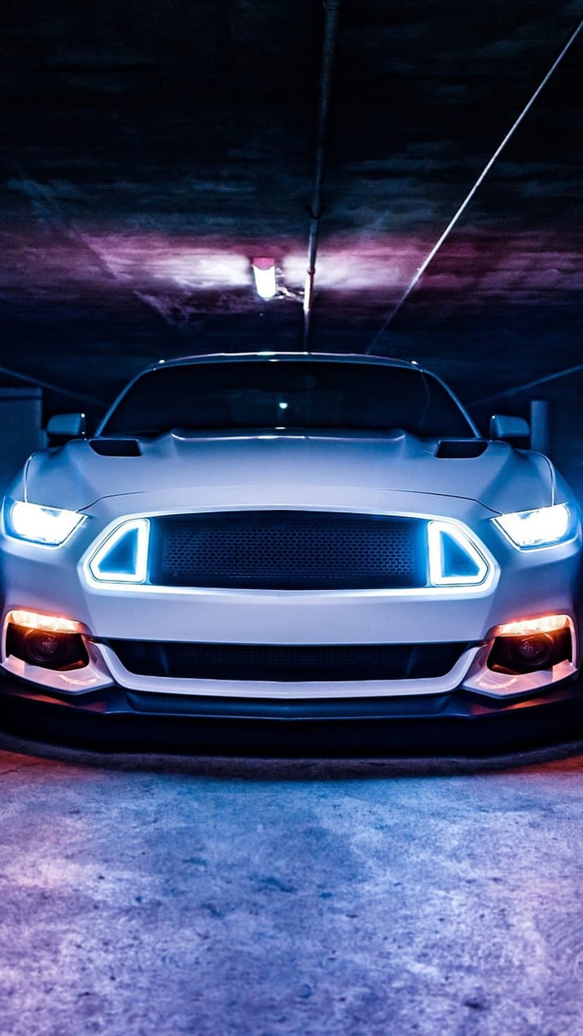 White Ford Mustang with blue neon headlight rings and orange bumper lights in a dark purple-lit parking garage.