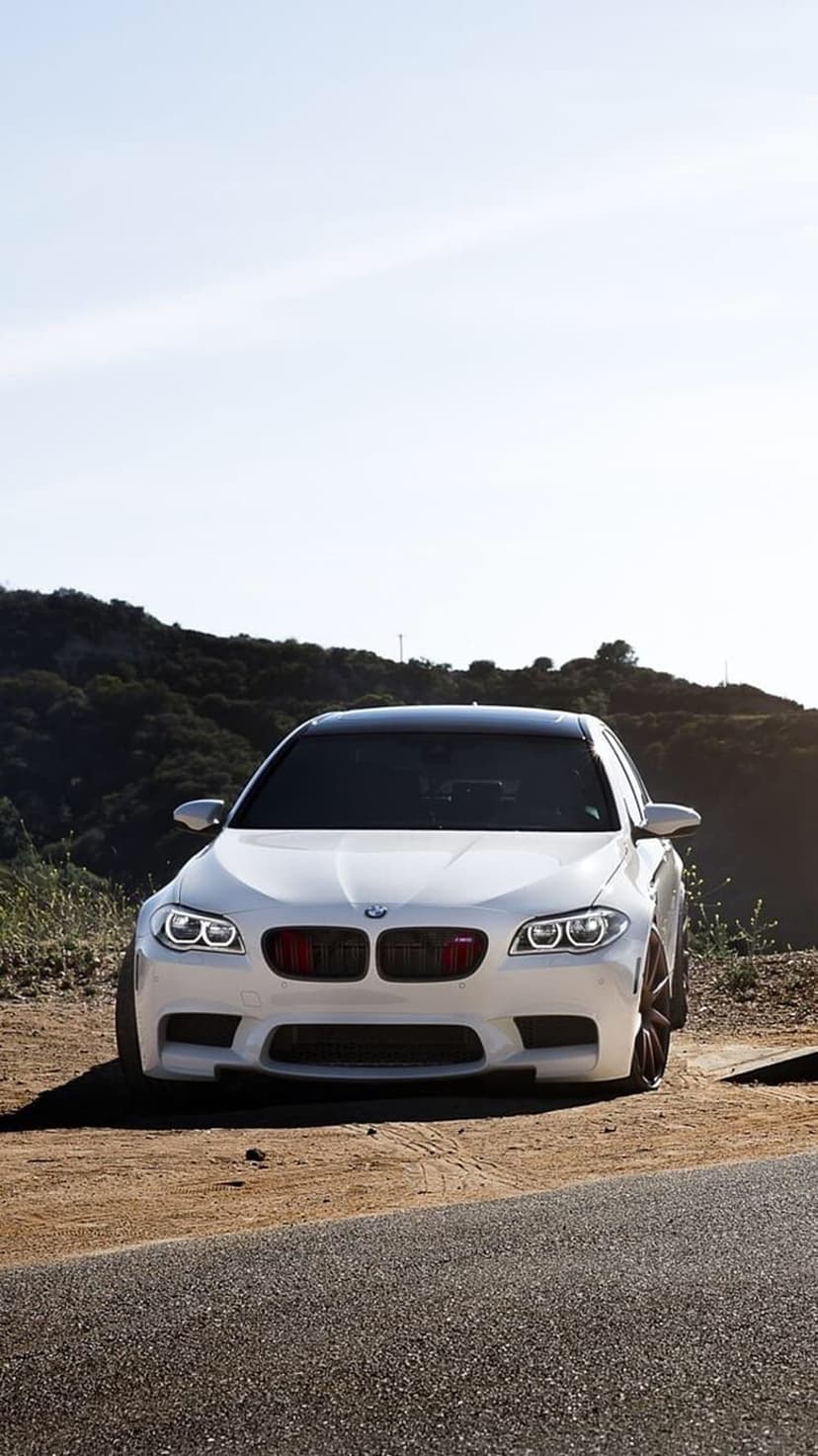 White BMW M5 sedan with red grille accents parked on a dirt road featuring lush green mountains in the distance.