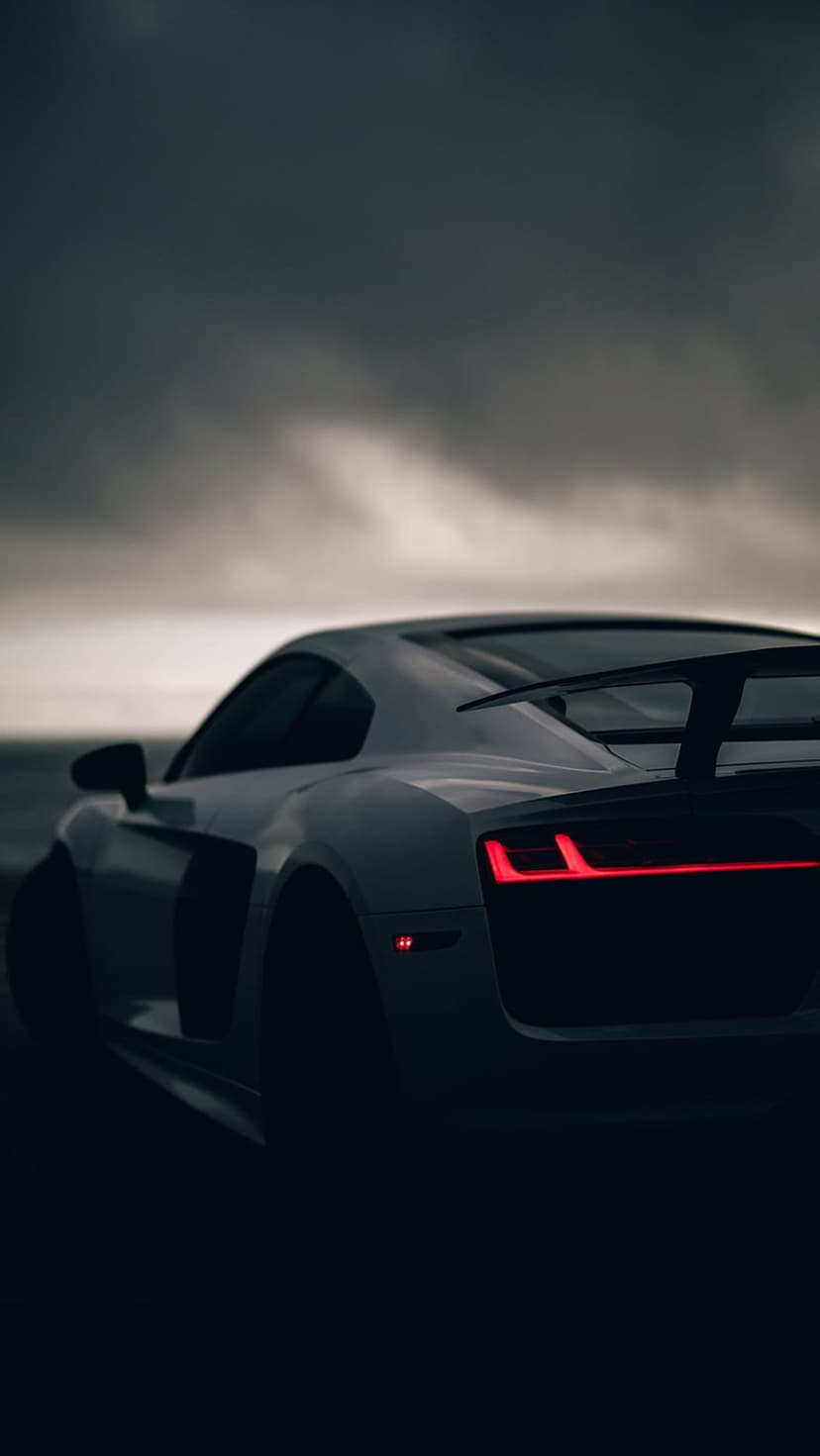 White sports car rear view with glowing red taillights parked by water under a cloudy evening sky at twilight.