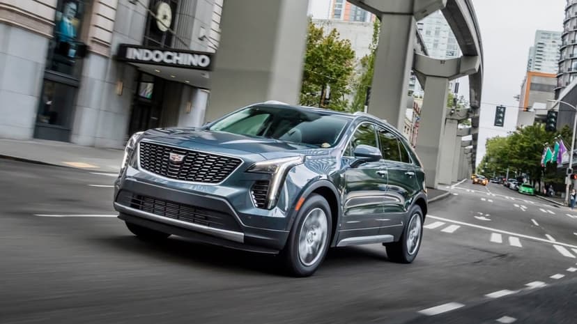 A metallic grey Cadillac XT4 SUV driving on a city street under elevated transit tracks near a storefront.