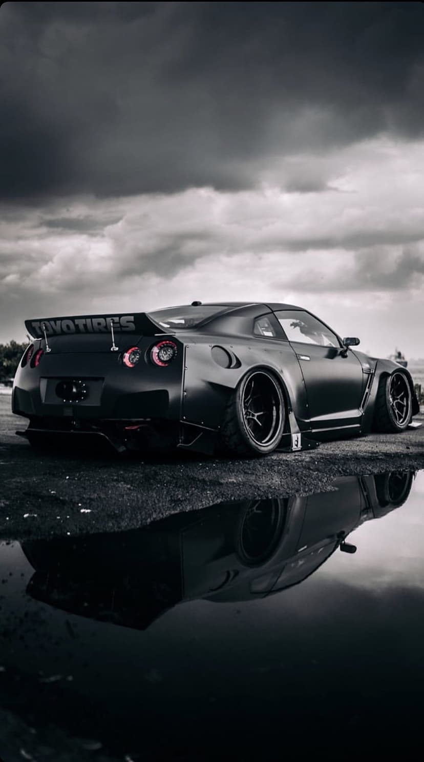 Matte black Nissan GT-R widebody car reflects in a street puddle beneath a dramatic and cloudy overcast sky.